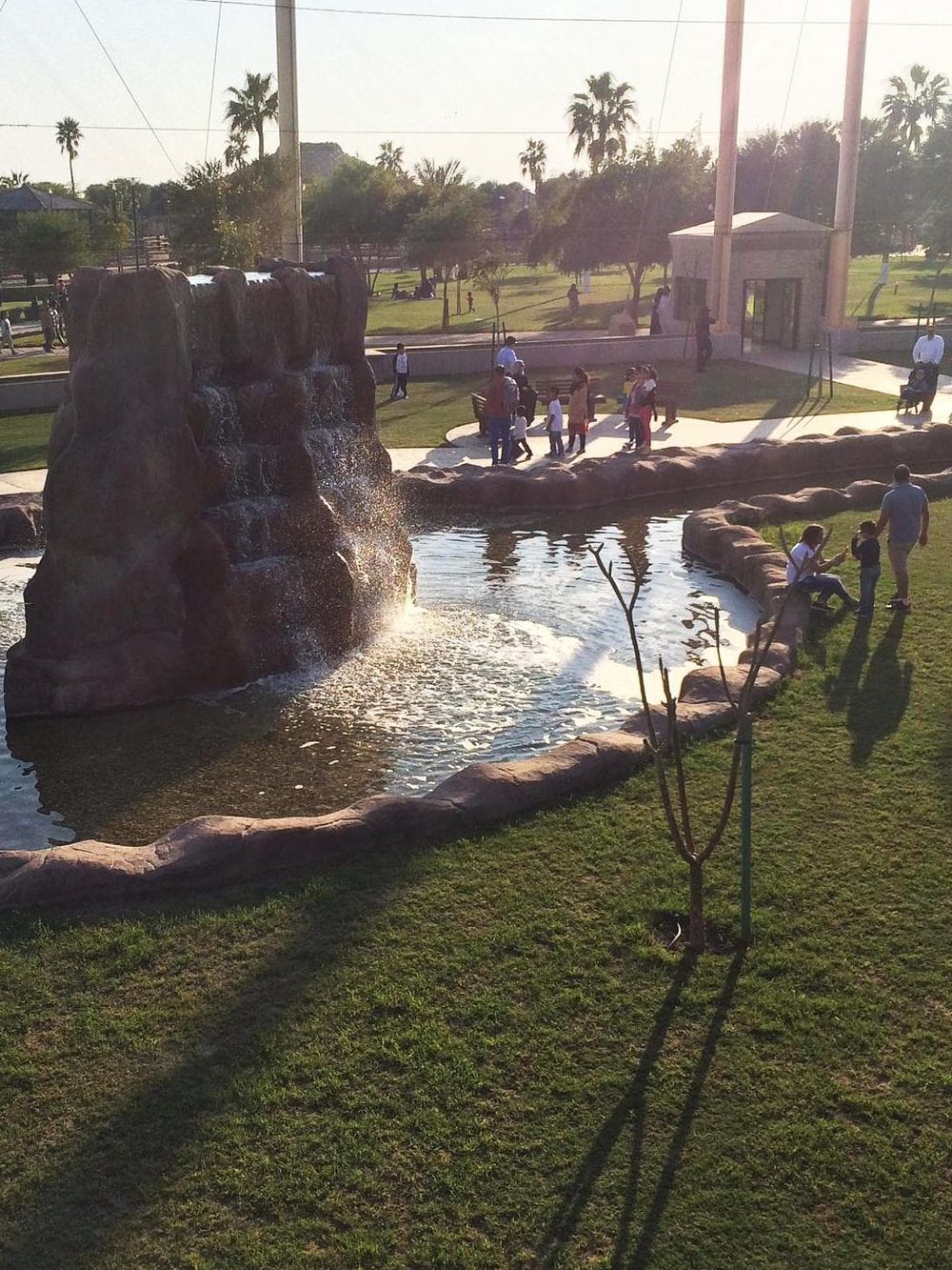 Water fountain with rocks in a park, surrounded by visitors, palm trees, and clear skies.