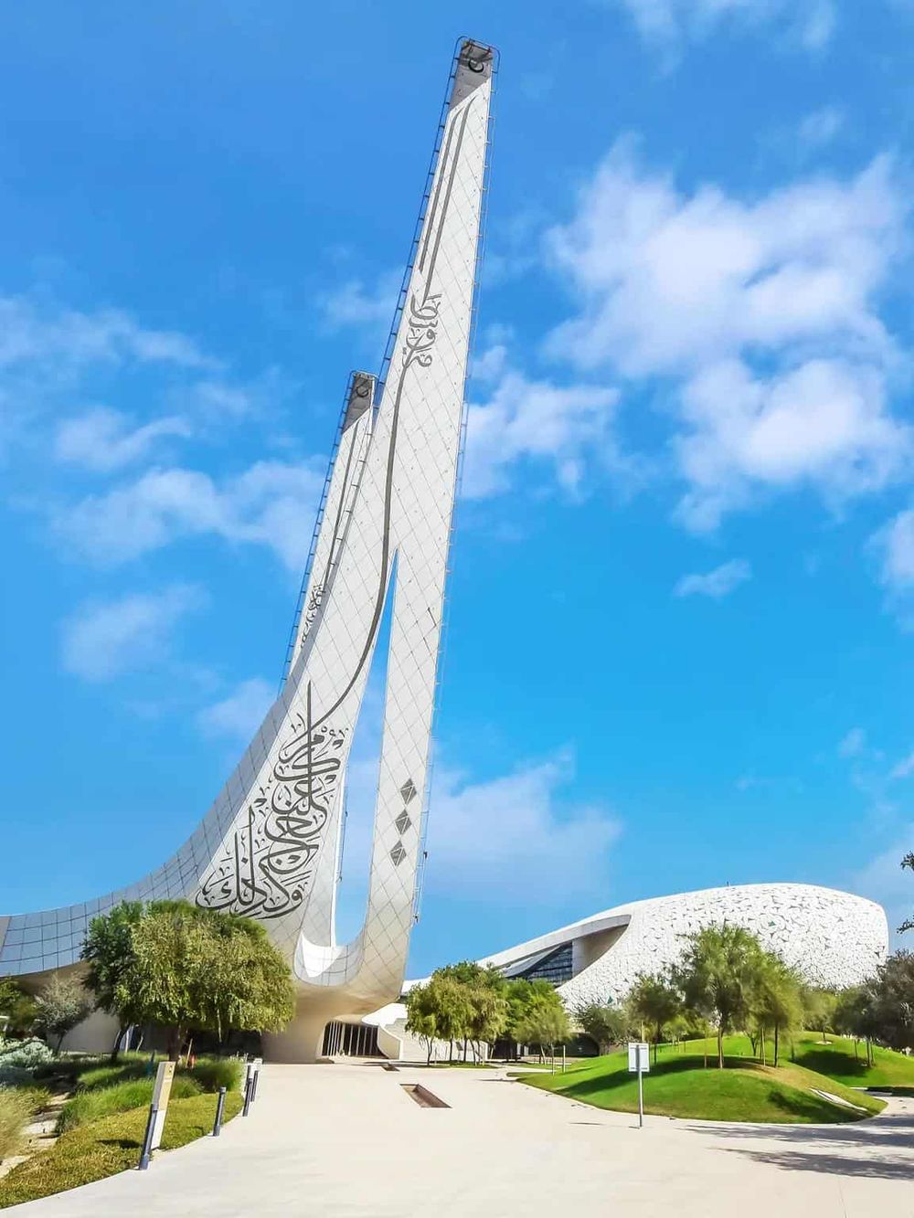 Modern mosque with tall minarets and contemporary architecture, surrounded by greenery and a clear blue sky.