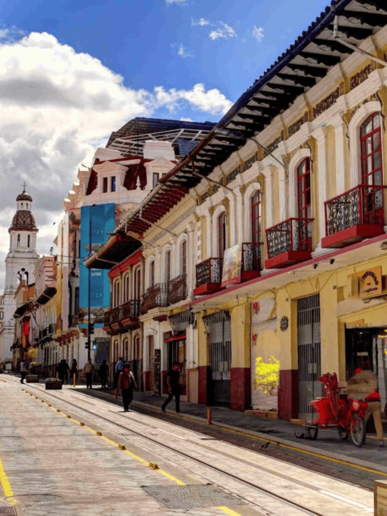 Colorful historic buildings on a lively street with trolley tracks, under a blue sky with clouds, in Queretaro, Mexico.