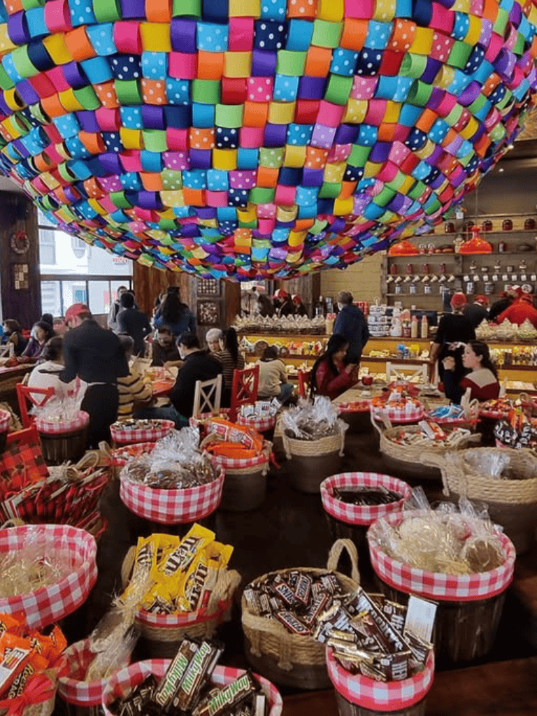 Colorful balloon ceiling overhead at festive holiday market with shoppers and gift baskets.