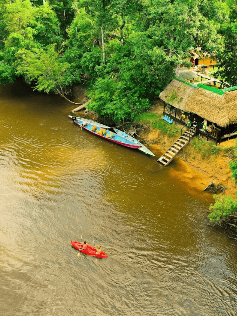Colorful boat docked along lush riverbank with kayaking scene nearby, scenic tropical setting.