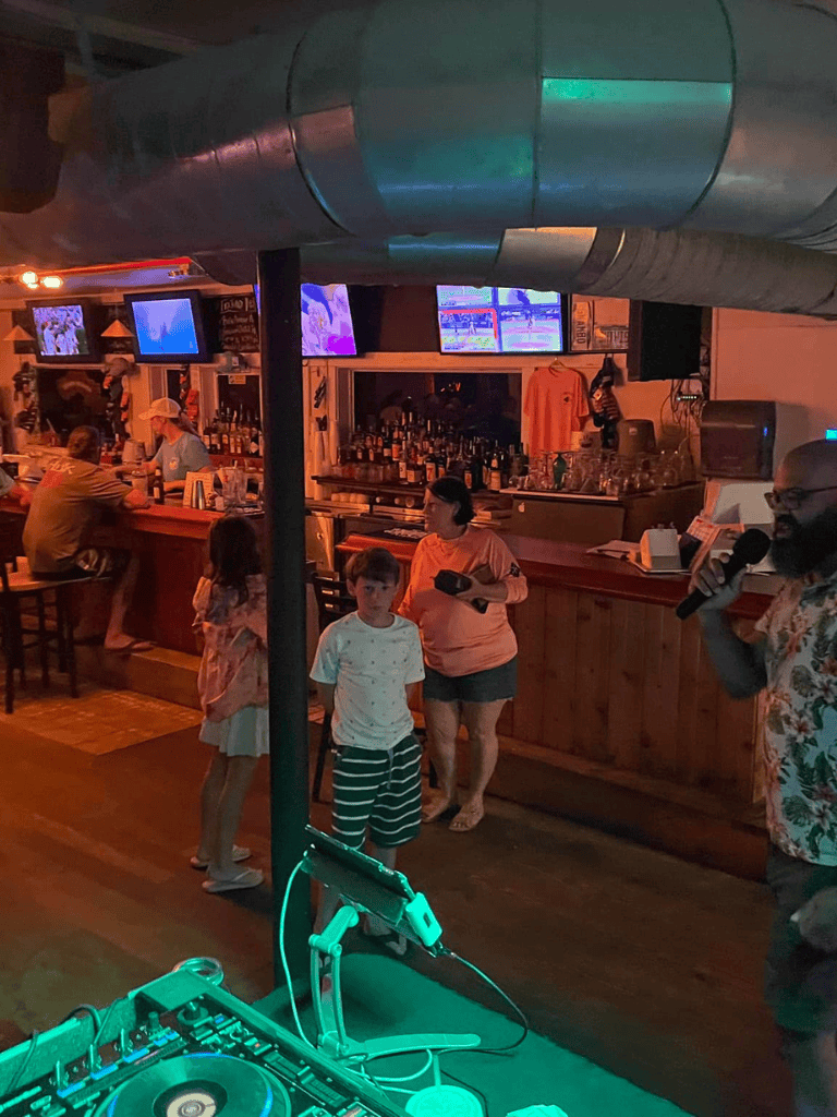 Crowd enjoying a live DJ at a bar, with children dancing, TVs showing sports, and colorful lighting ambiance.