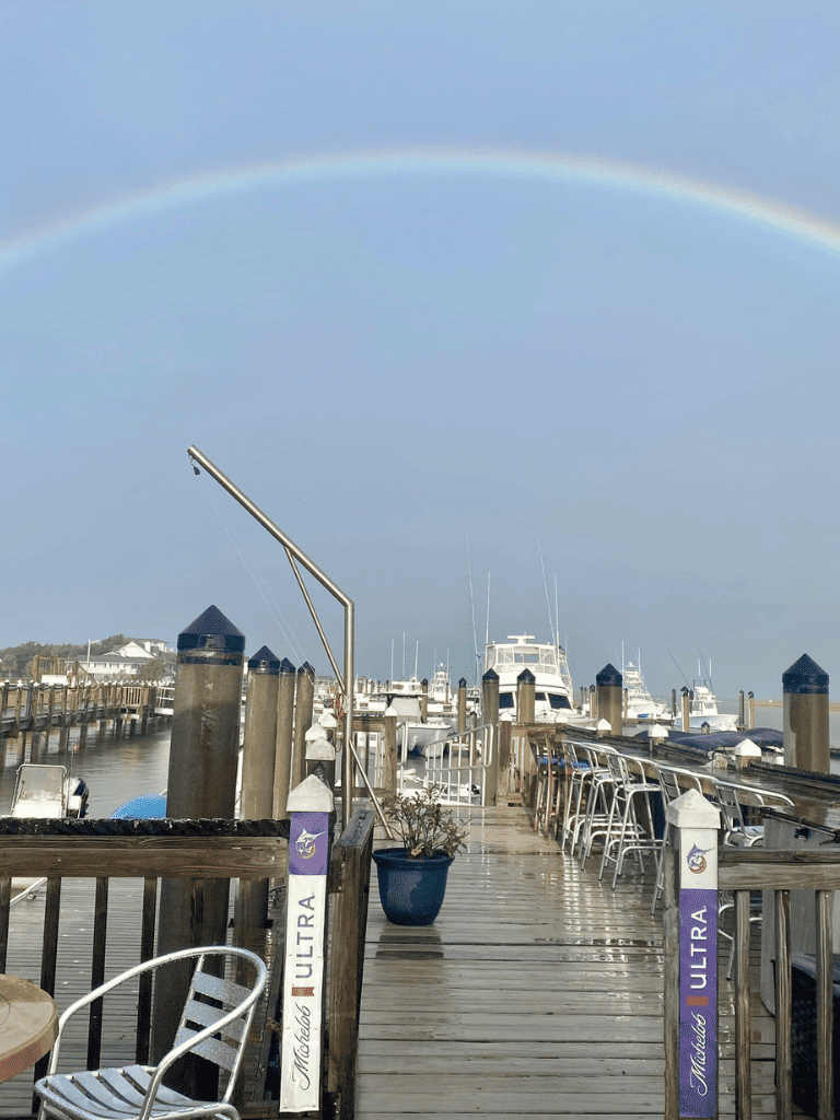Bright rainbow over marina with boats and dock, showcasing scenic waterfront views and cloudy sky.