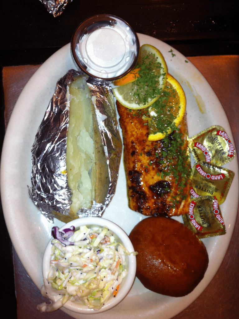 Crispy baked salmon with lemon and herbs, served with a baked potato, coleslaw, and a corned beef ball, on a white plate.