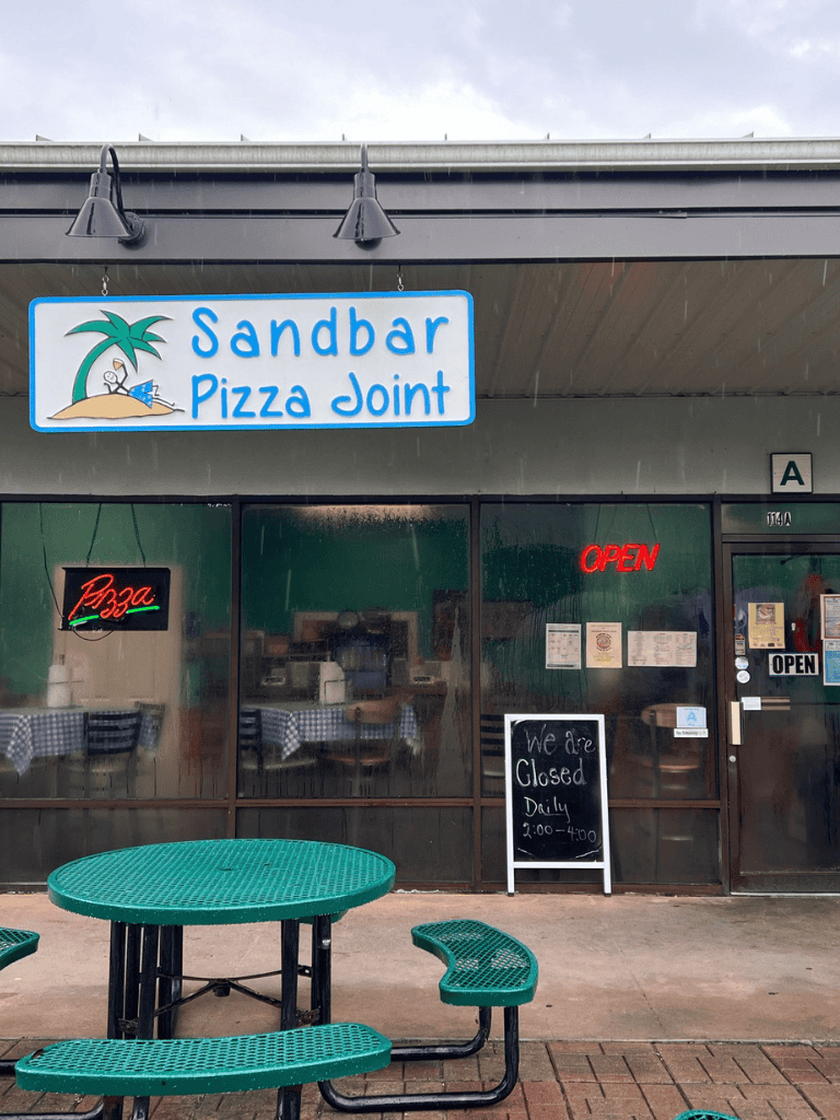 Sandbar Pizza Joint restaurant on rainy day, outdoor seating available, sign shows daily hours.