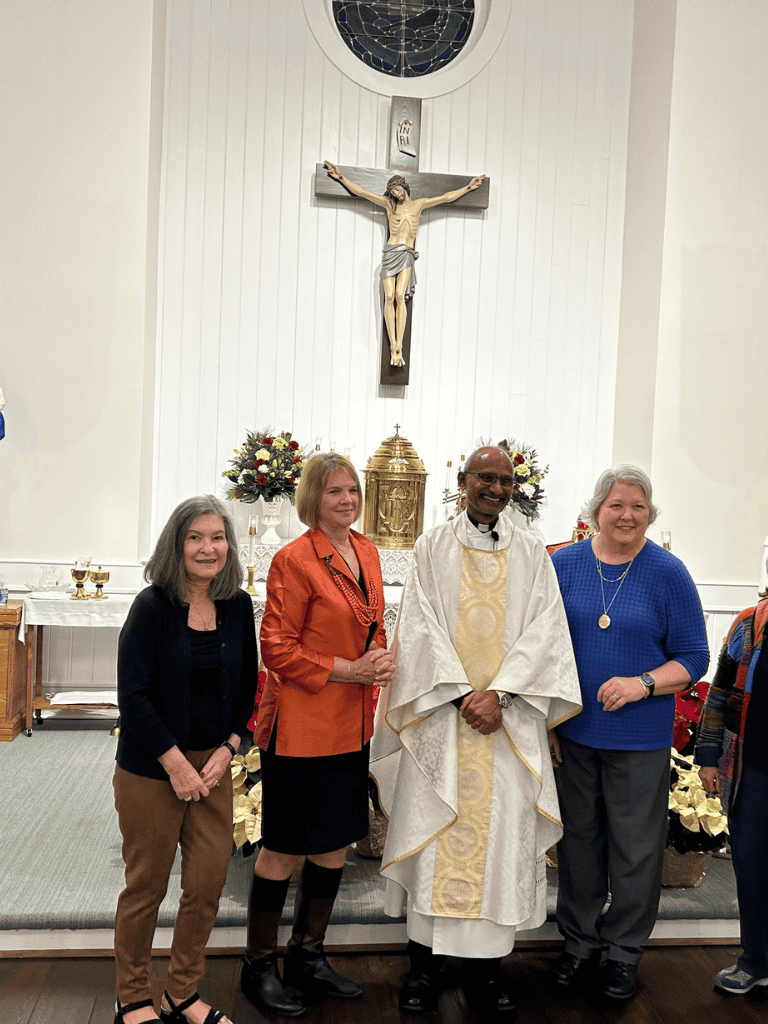 Peaceful church interior with a priest and three women standing in front of a crucifix and altar.
