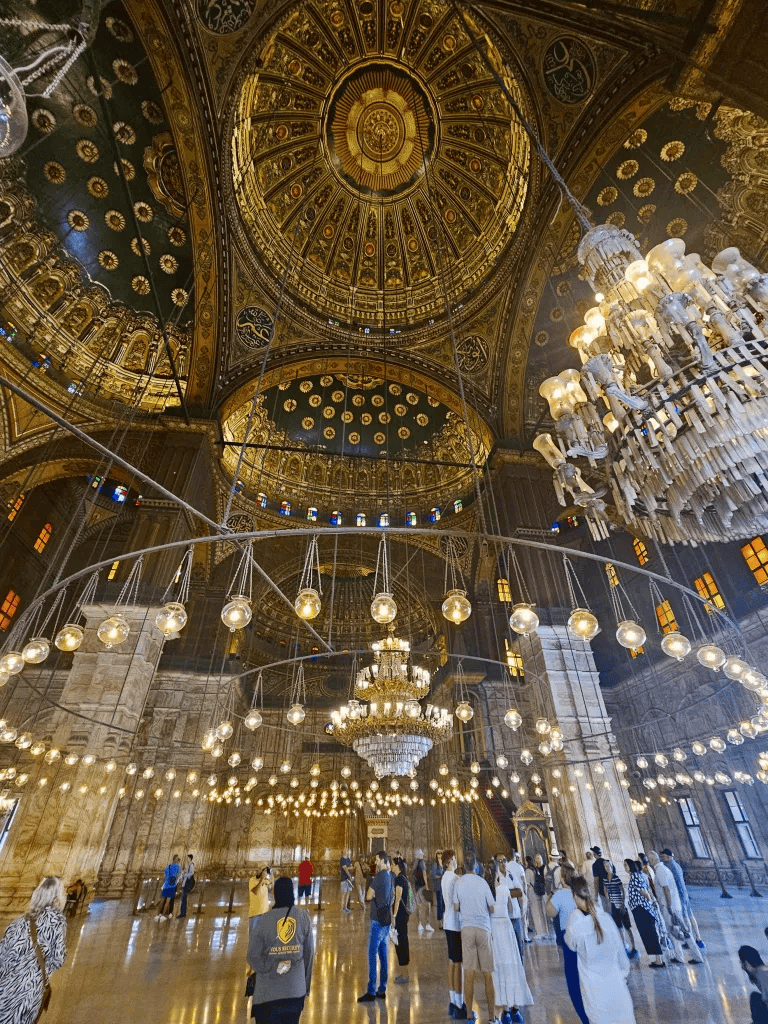 Intricate gold and crystal chandeliers in a grandhistoric mosque dome with visitors exploring the interior.
