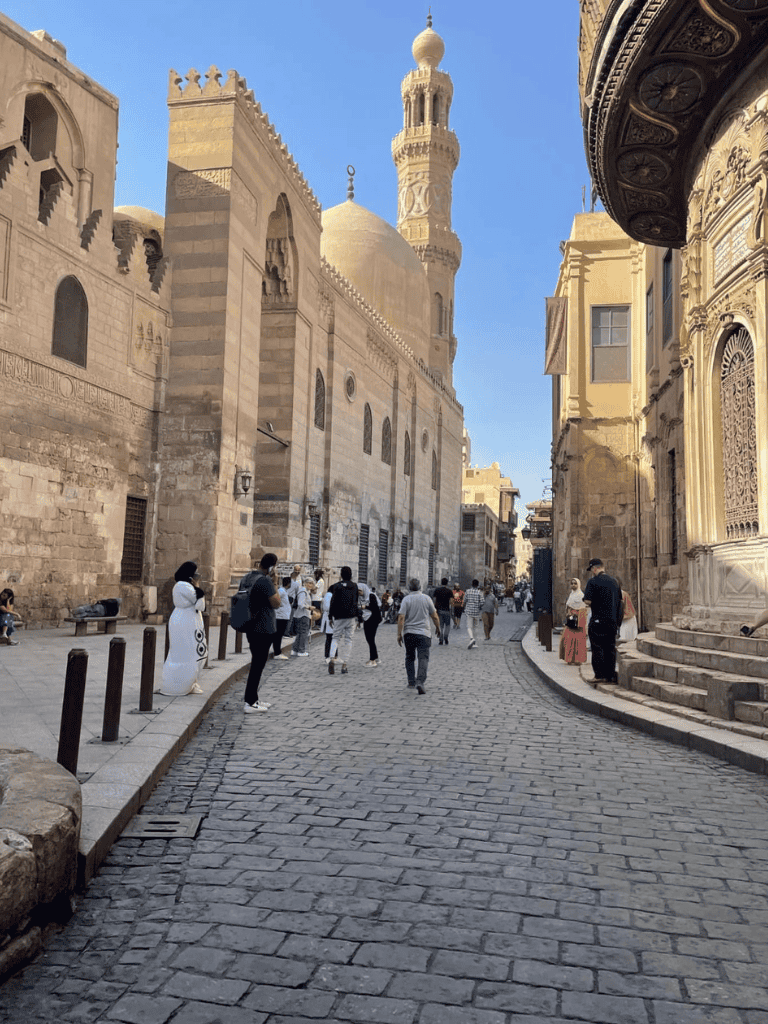 Quiet street in Cairo near a historic mosque and ornate buildings, bustling with people and tourists.