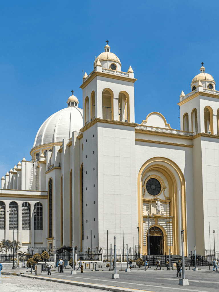 Imposing white church with domes and bell towers against clear blue sky, street scene with pedestrians.