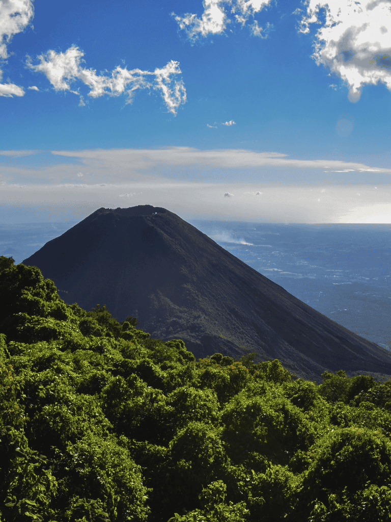 Lush green forest leading to a volcanic mountain under a blue sky with clouds.