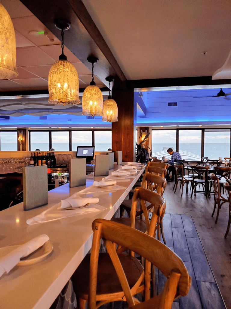 Cozy seaside restaurant interior with ocean view, warm lighting, and stylish wooden furniture.