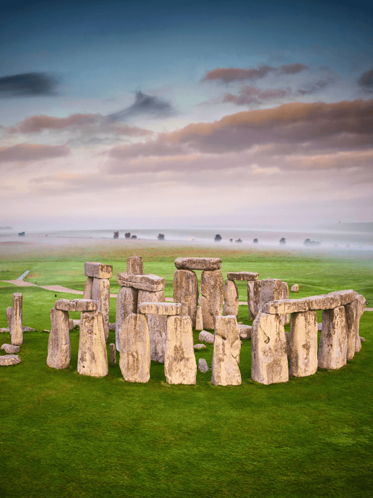 Ancient Stonehenge monument at sunrise with cloudy sky and green landscape in the background.