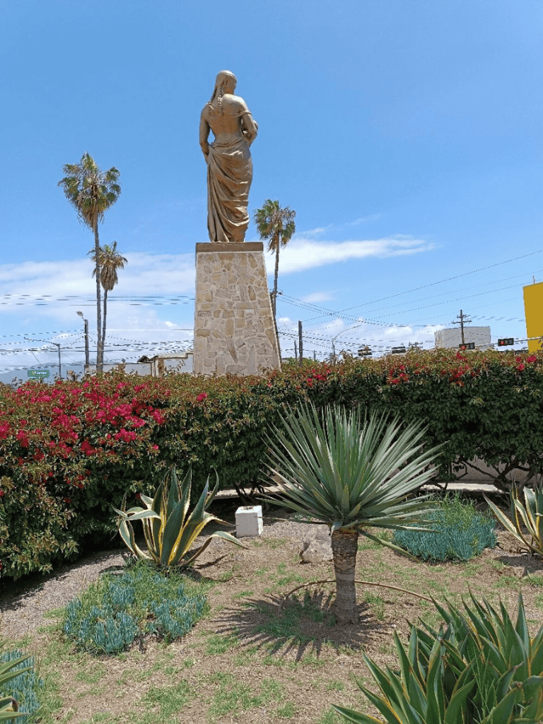 Ancient statue with blooming desert plants and palm trees under a bright blue sky.