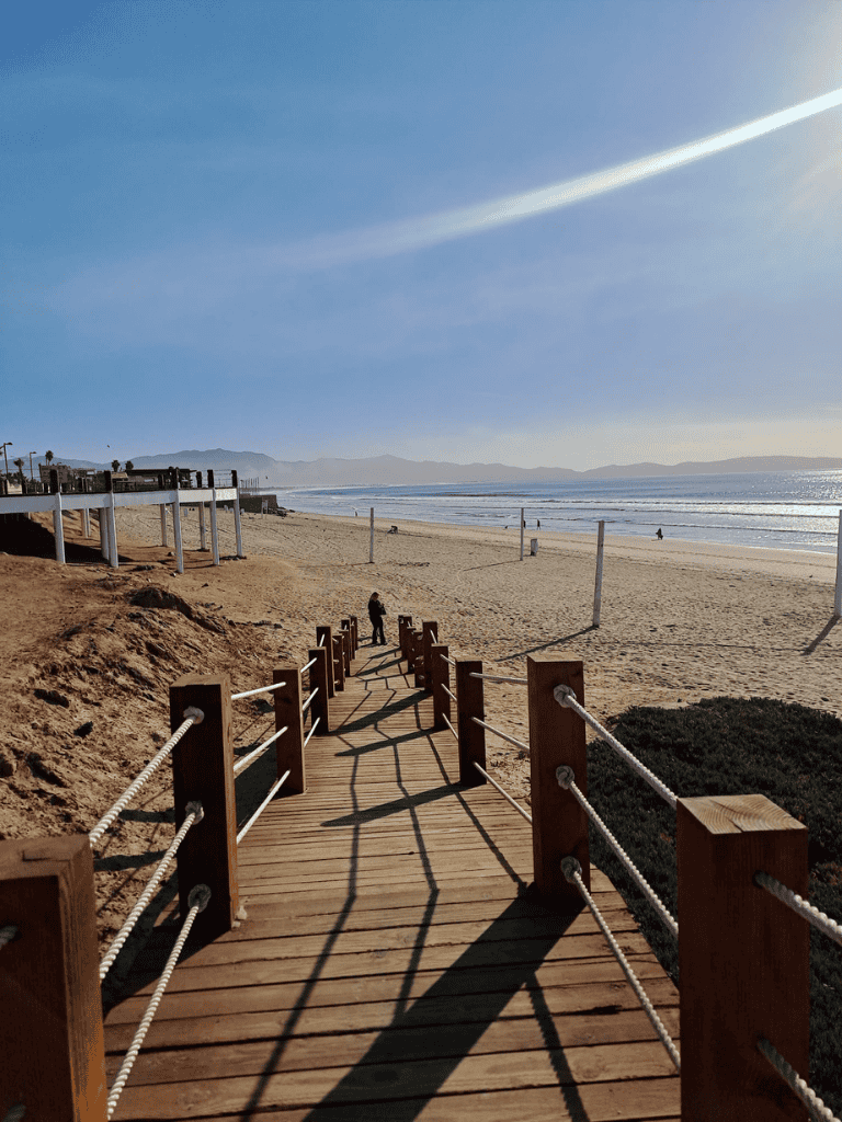 Bright wooden pathway leading to sunny beach with ocean view and clear sky.