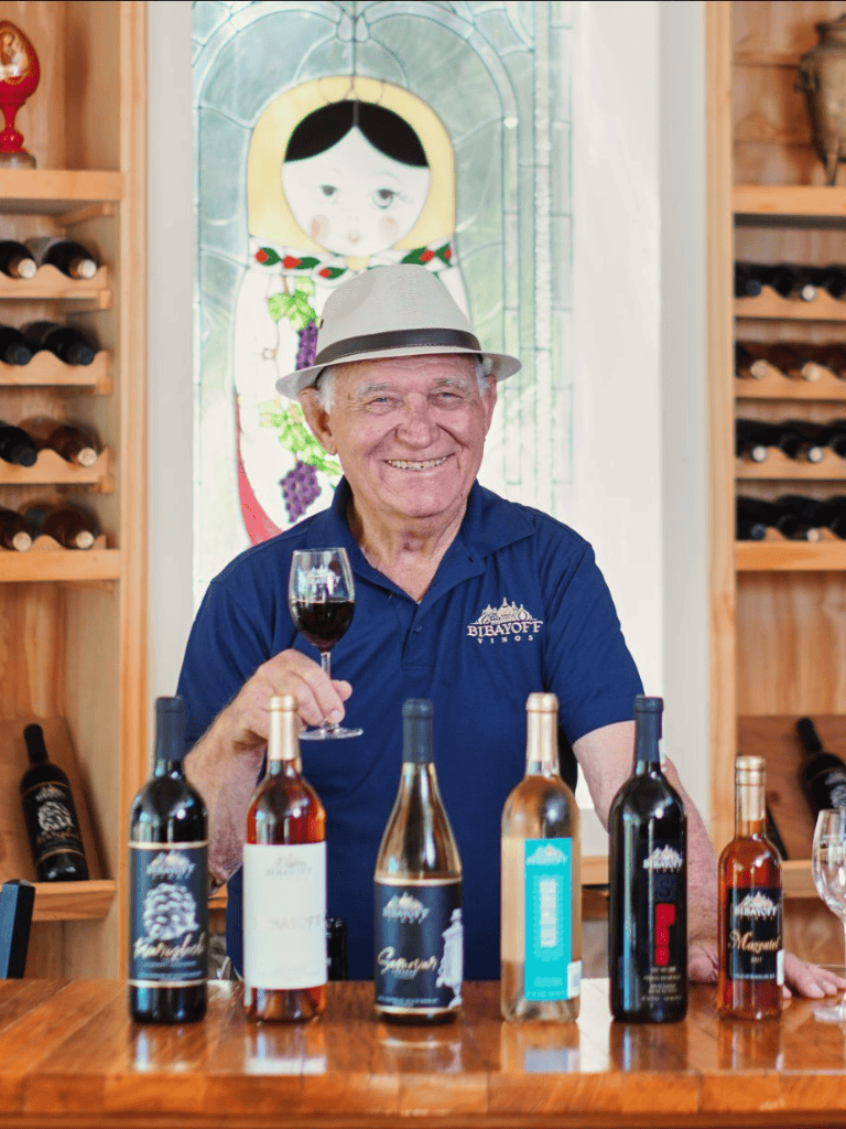 Aged man tasting wine in winery with bottles and colorful artwork in background.