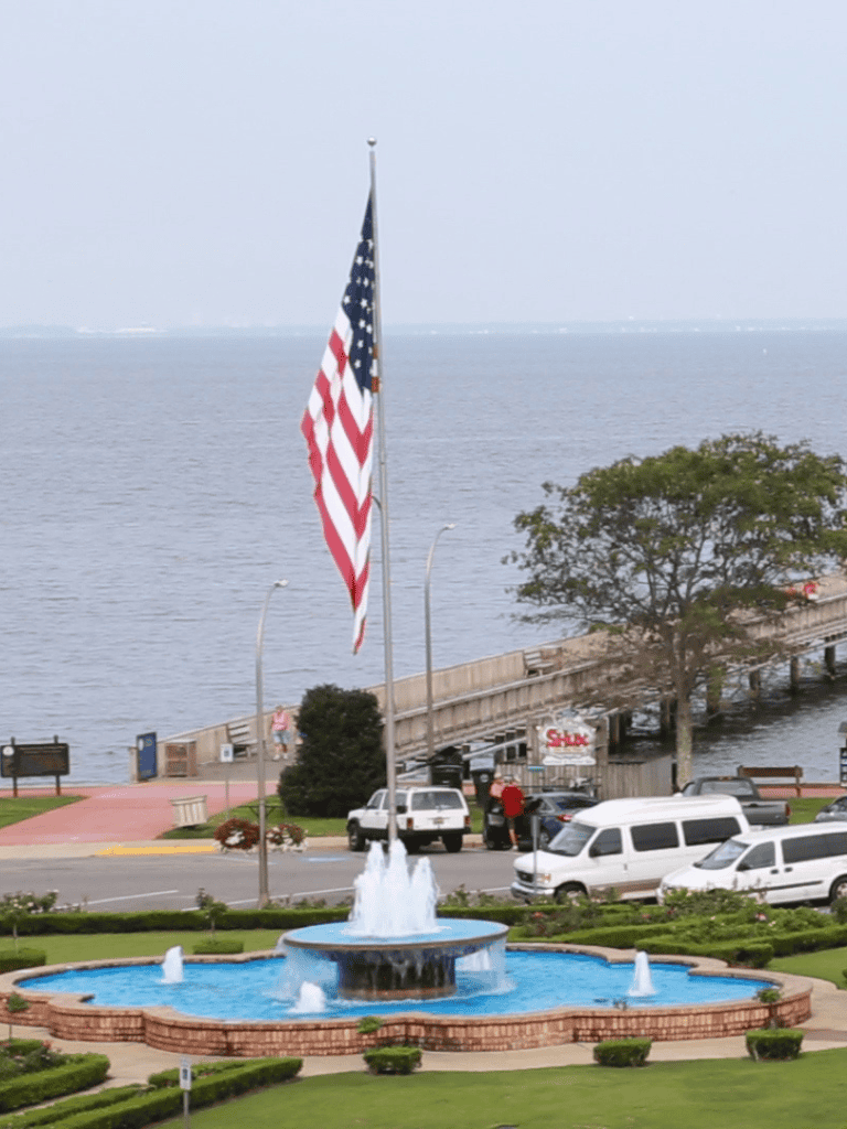 American flag at waterfront park with fountain and parking area, scenic ocean view, and downtown skyline in background.