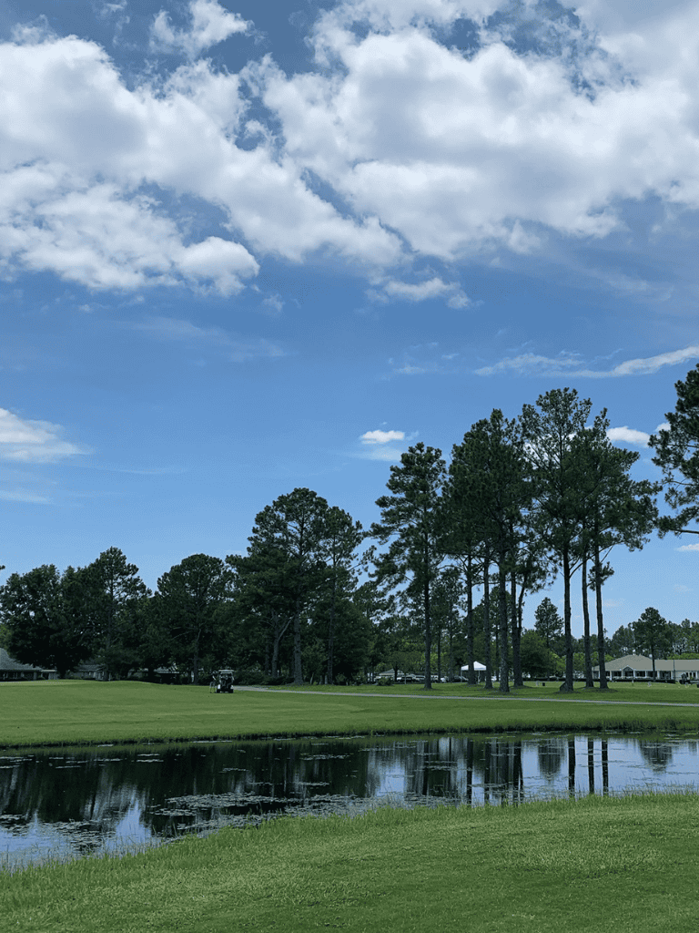 Lush golf course with water hazard and tall pine trees under a partly cloudy sky.