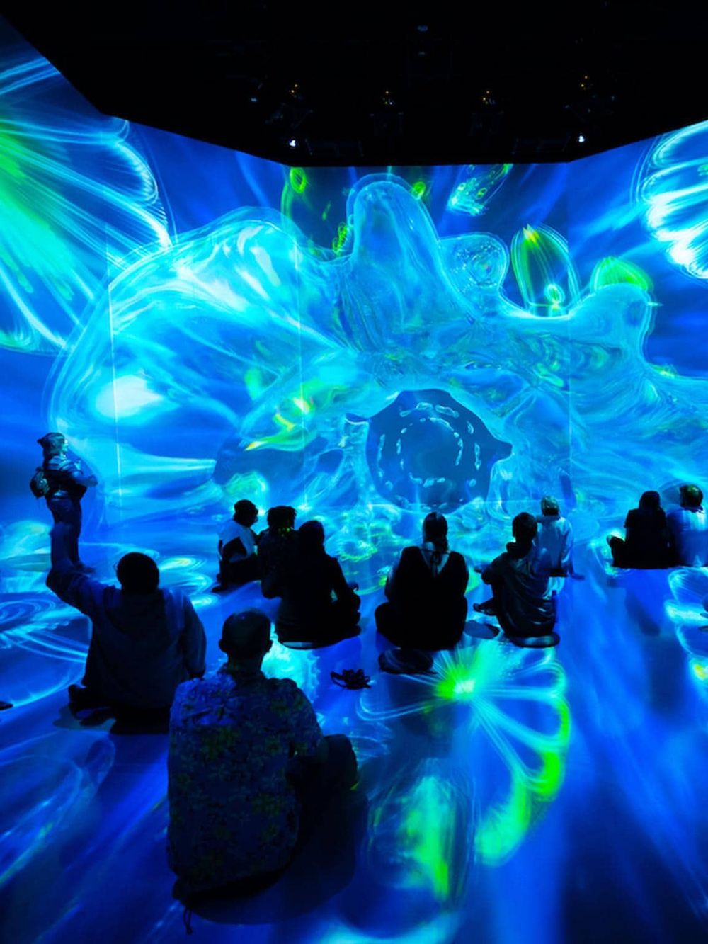 Vivid digital art installation with visitors seated on the floor, immersed in colorful, swirling blue, green, and yellow lights.