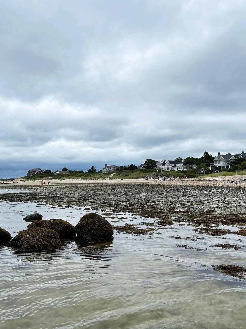 Expansive beach scene with rocks, sand, and ocean, under cloudy sky with beachfront homes in the background.