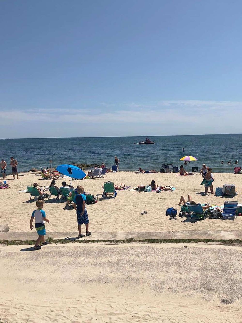 Kids playing on a sandy beach with umbrellas, sunbathers, and a boat in the ocean.