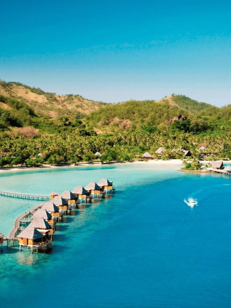 Overwater bungalows on tropical island with turquoise water and lush green hills in background.