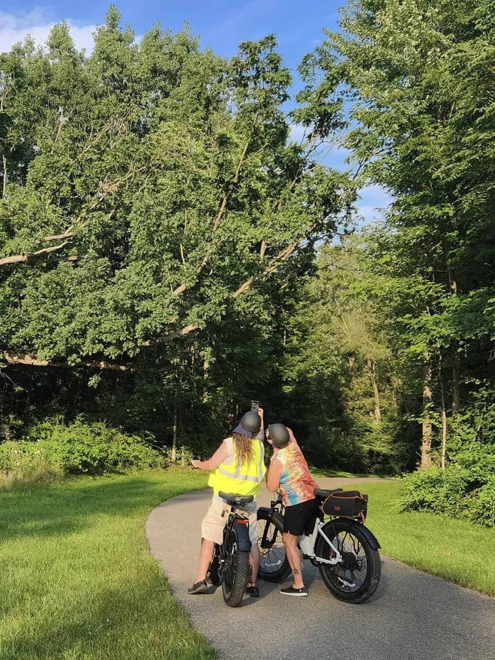 Two women with bikes exploring nature on a scenic trail with lush green trees and clear blue sky.