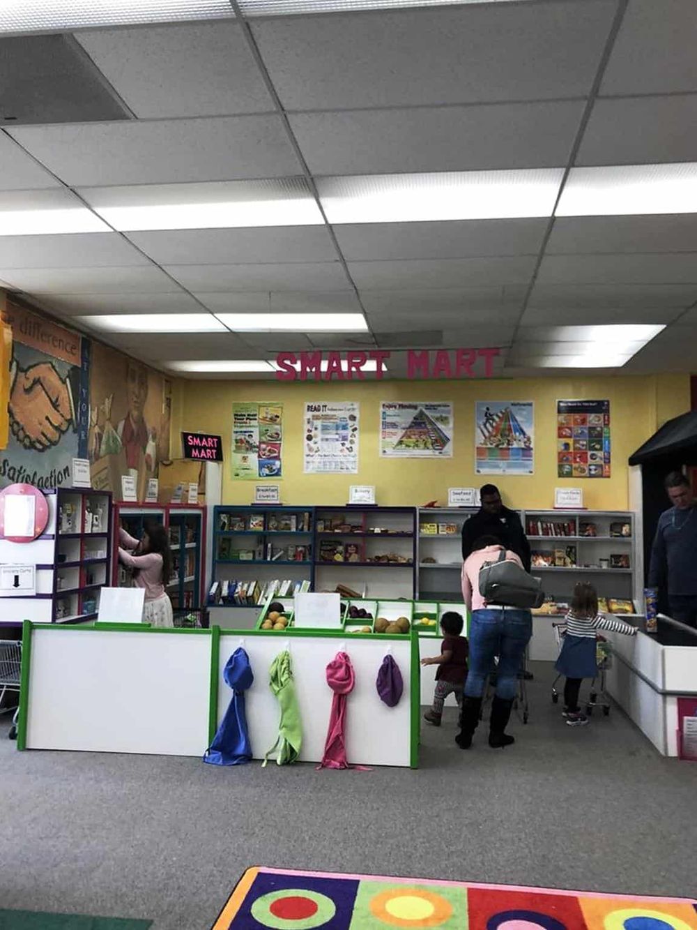 Bright indoor retail store with children and adults shopping at a market-style kiosk, featuring "Smart Mart" signage.