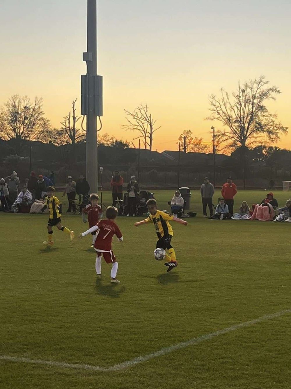 Young children playing soccer on a field during sunset, surrounded by spectators.