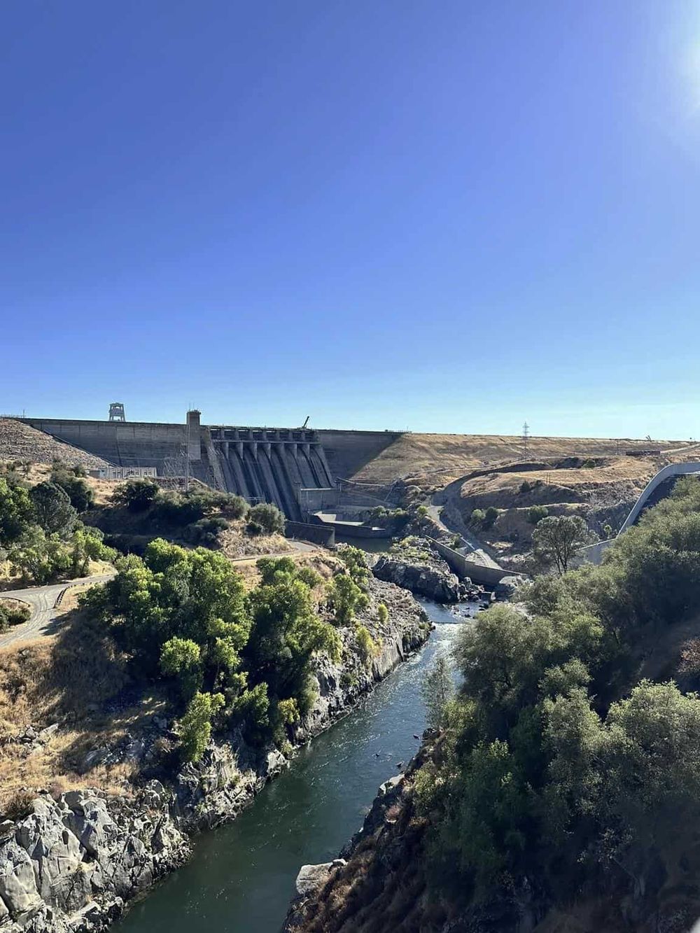Dam and river landscape in California with clear blue sky, lush greenery, and rugged terrain.