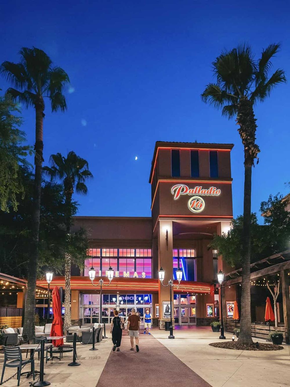 Brightly lit Palladio 16 Cinema at dusk with palm trees, marquee, and twilight sky.