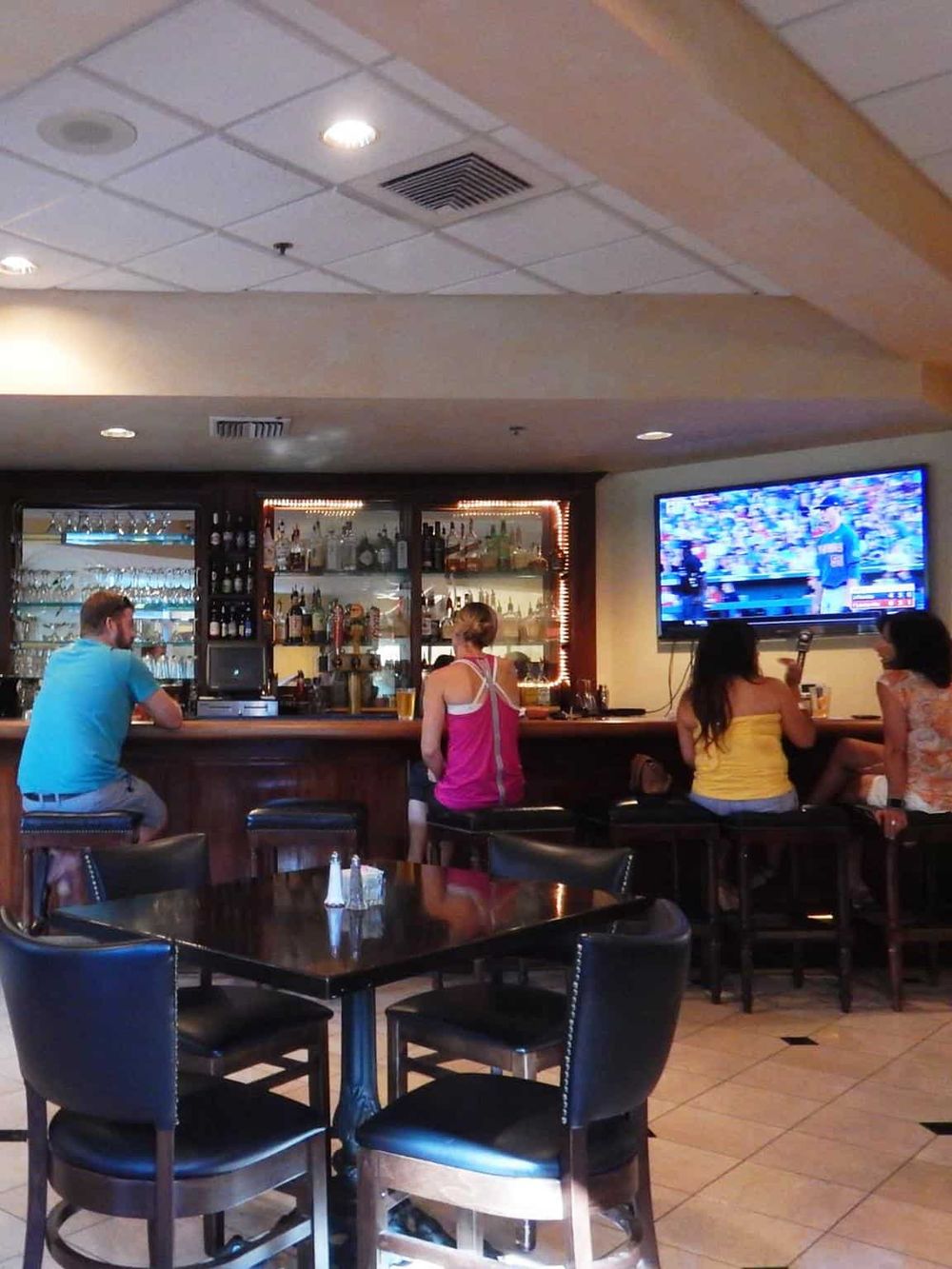 Comfortable bar with patrons watching sports, featuring a TV screen, bar counter, and seating area for socializing and dining.