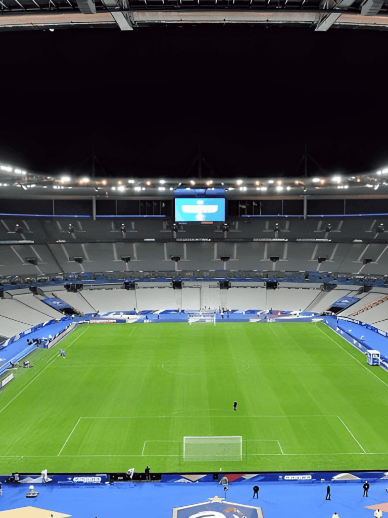 Brightly lit soccer stadium with green pitch, empty seats, and large screen for fans and players.