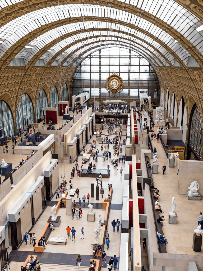 Impressive grand hall of the Musée d'Orsay with high glass vaulted ceilings and classical sculptures, bustling with visitors.