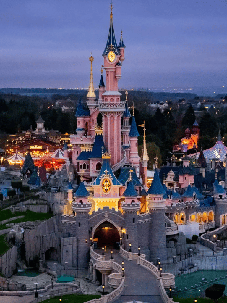 Pink Disneyland castle at dusk with illuminated spires and surrounding themed attractions.