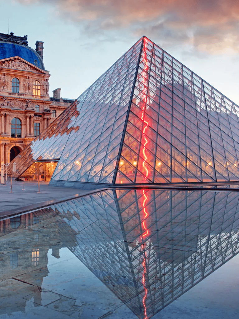 Modern glass pyramid entrance at the Louvre Museum in Paris with reflective water pool, illuminated by sunset lighting.