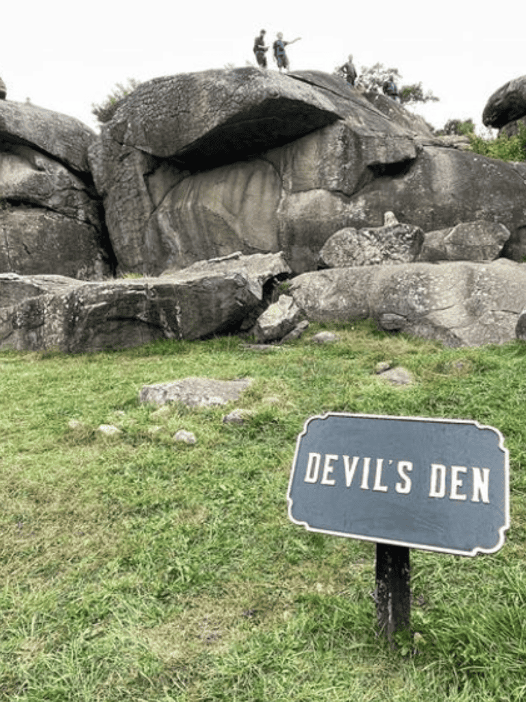 Dark rock formation called Devil's Den with a sign in front, outdoor scenic location, popular for hiking and exploring.