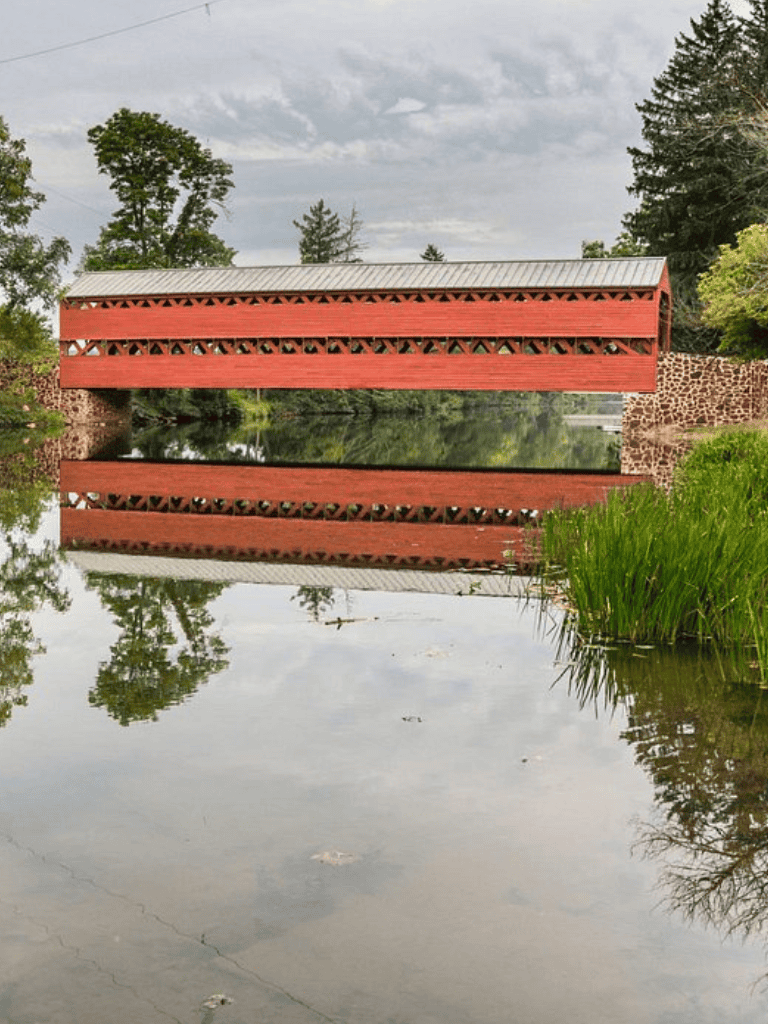 Colorful covered bridge over calm water with reflections, surrounded by lush greenery and trees.