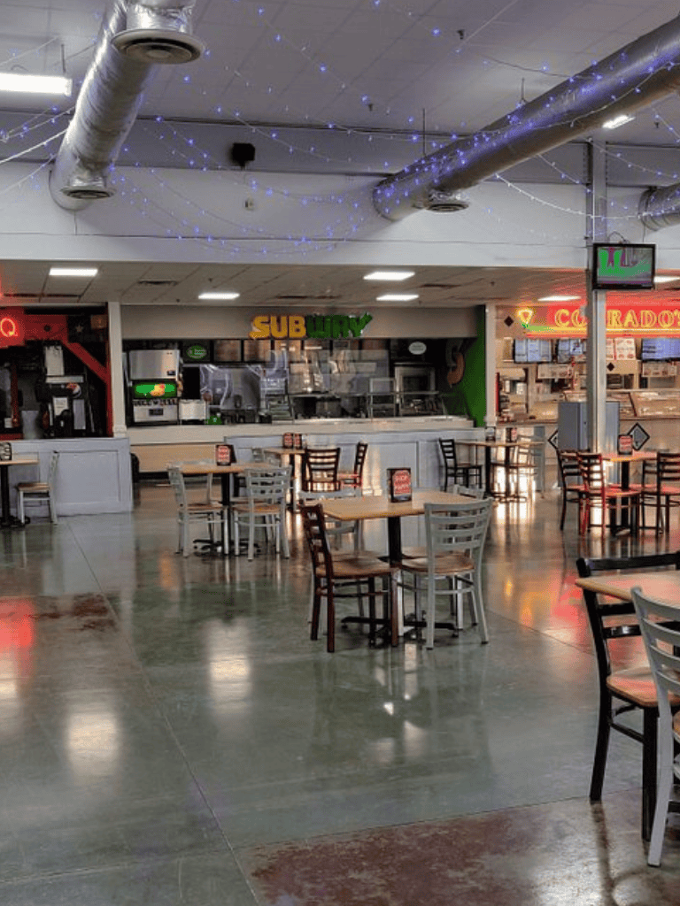 Empty food court with Subway restaurant and other eateries in the background, decorated with blue string lights and industrial ceiling ducts.