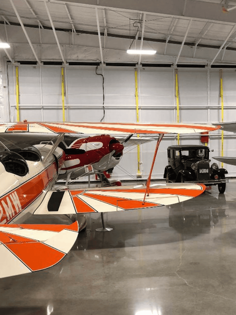 Small aircraft inside aviation museum with vintage vehicle in the background.