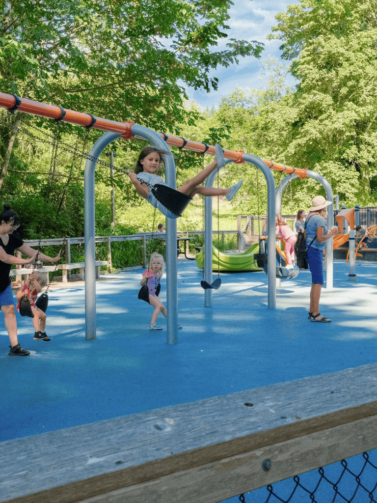 Swinging children at outdoor playground with lush green trees, family fun activities, and safe play area.