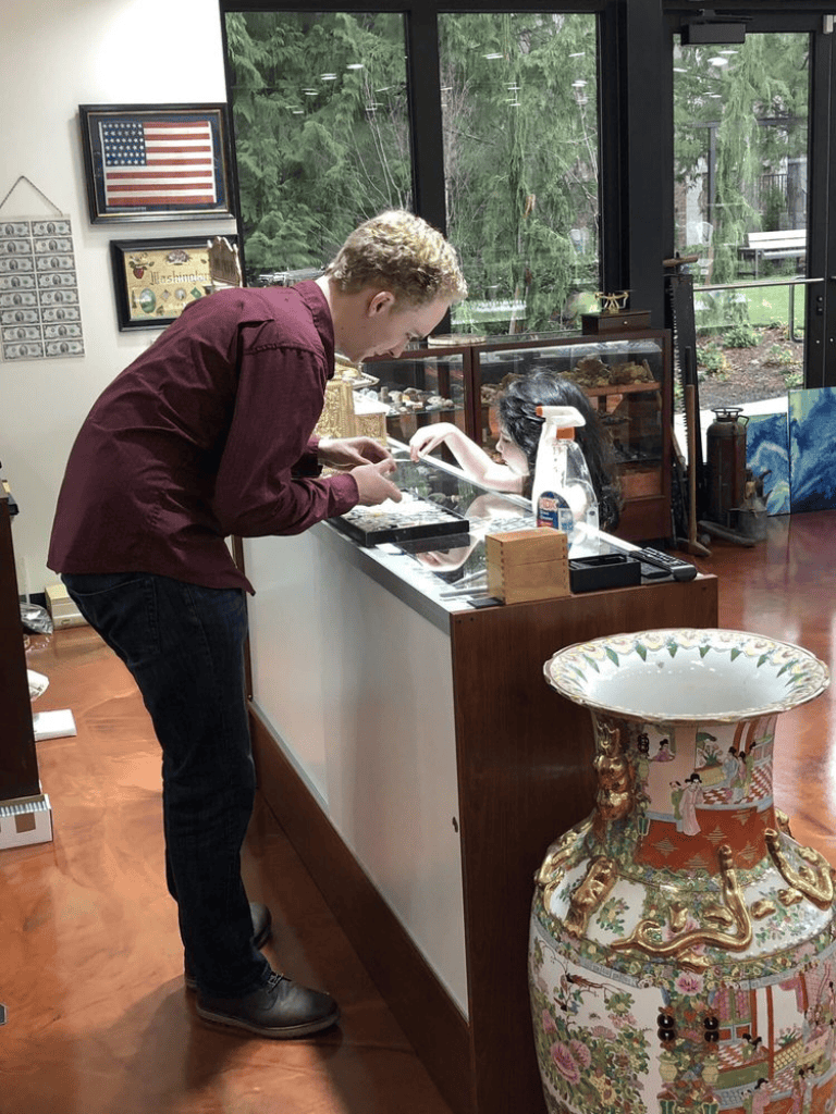 Man examining jewelry at antique shop with pottery and display case in background.