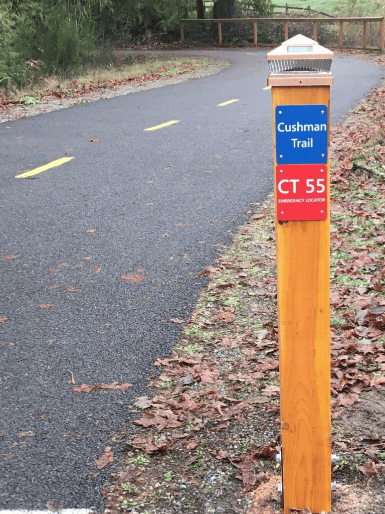 Cushman Trail directional sign with emergency locator, scenic paved path for walking and biking.