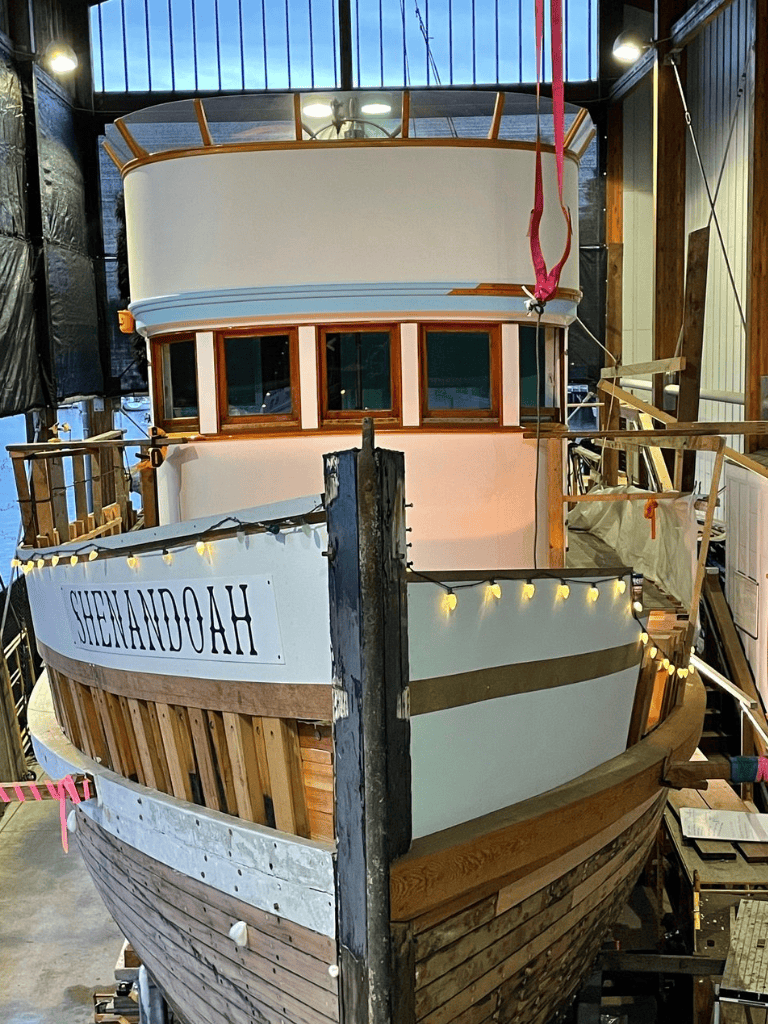 Boat construction in progress at a workshop, showcasing plank and frame assembly, boat name "SHENANDOAH" visible.