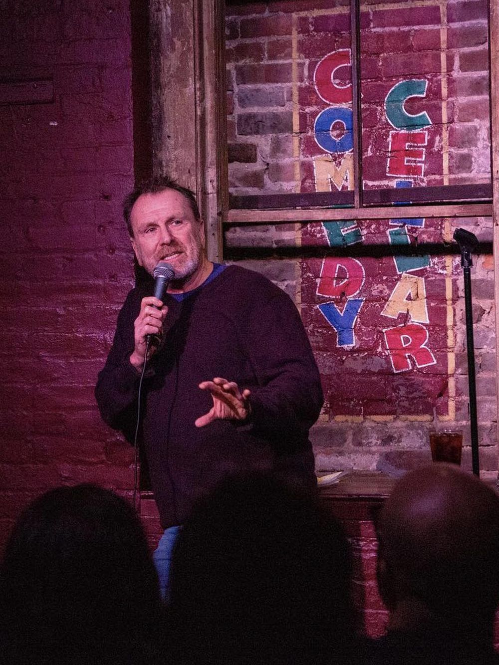 Warm comedy club scene with a male comedian performing with a microphone at Quest for Directions.