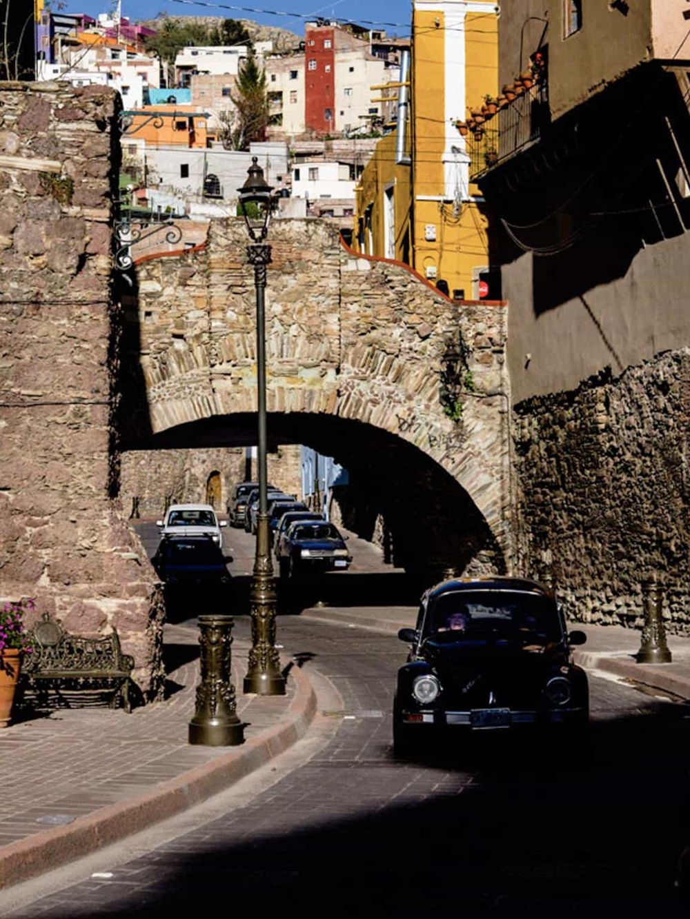 Colorful hillside neighborhood with historic stone bridge and vintage car in Mexico City.