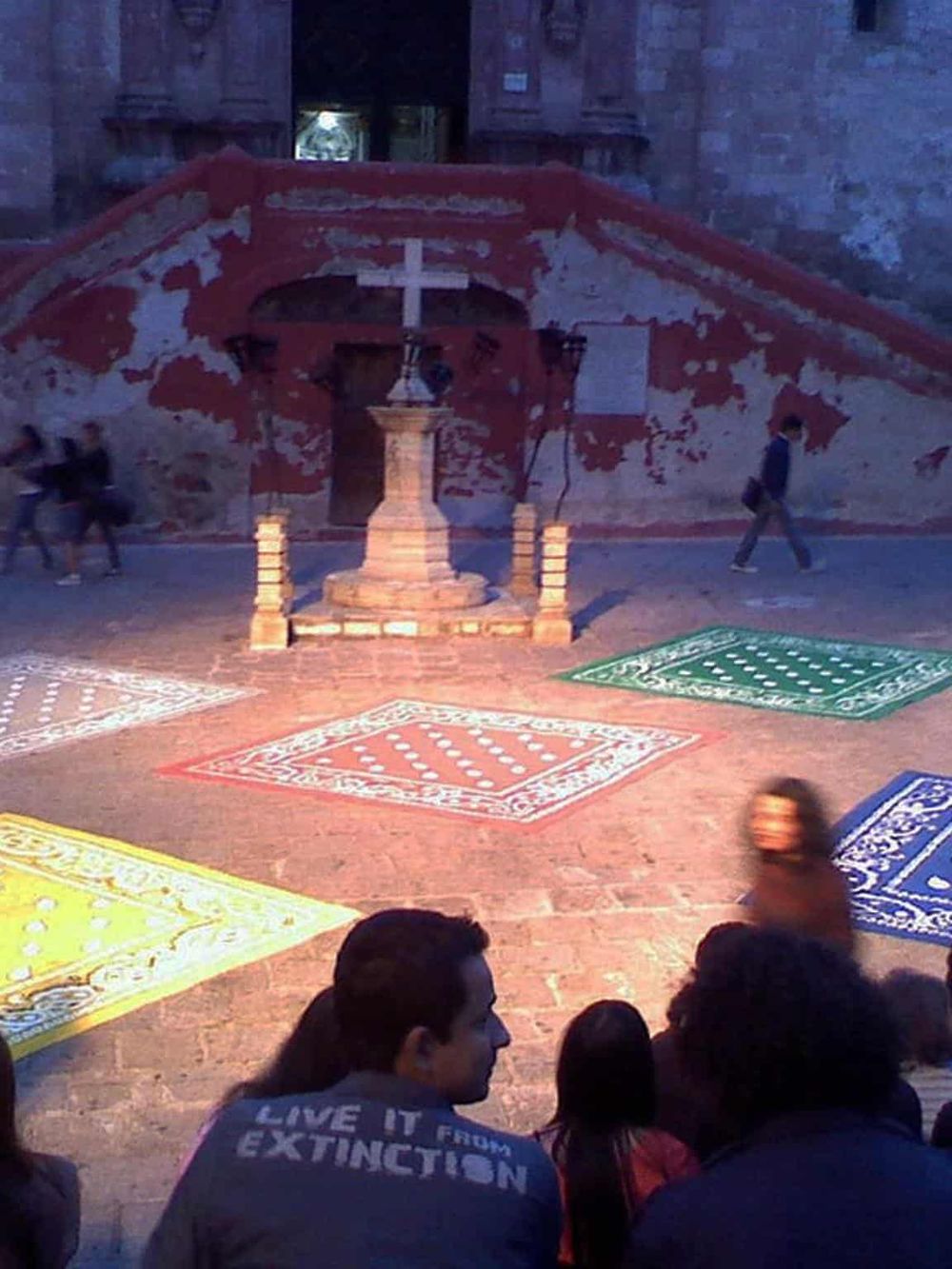 Colorful patterned rugs on cobblestone street with religious monument and historic brick wall in background.