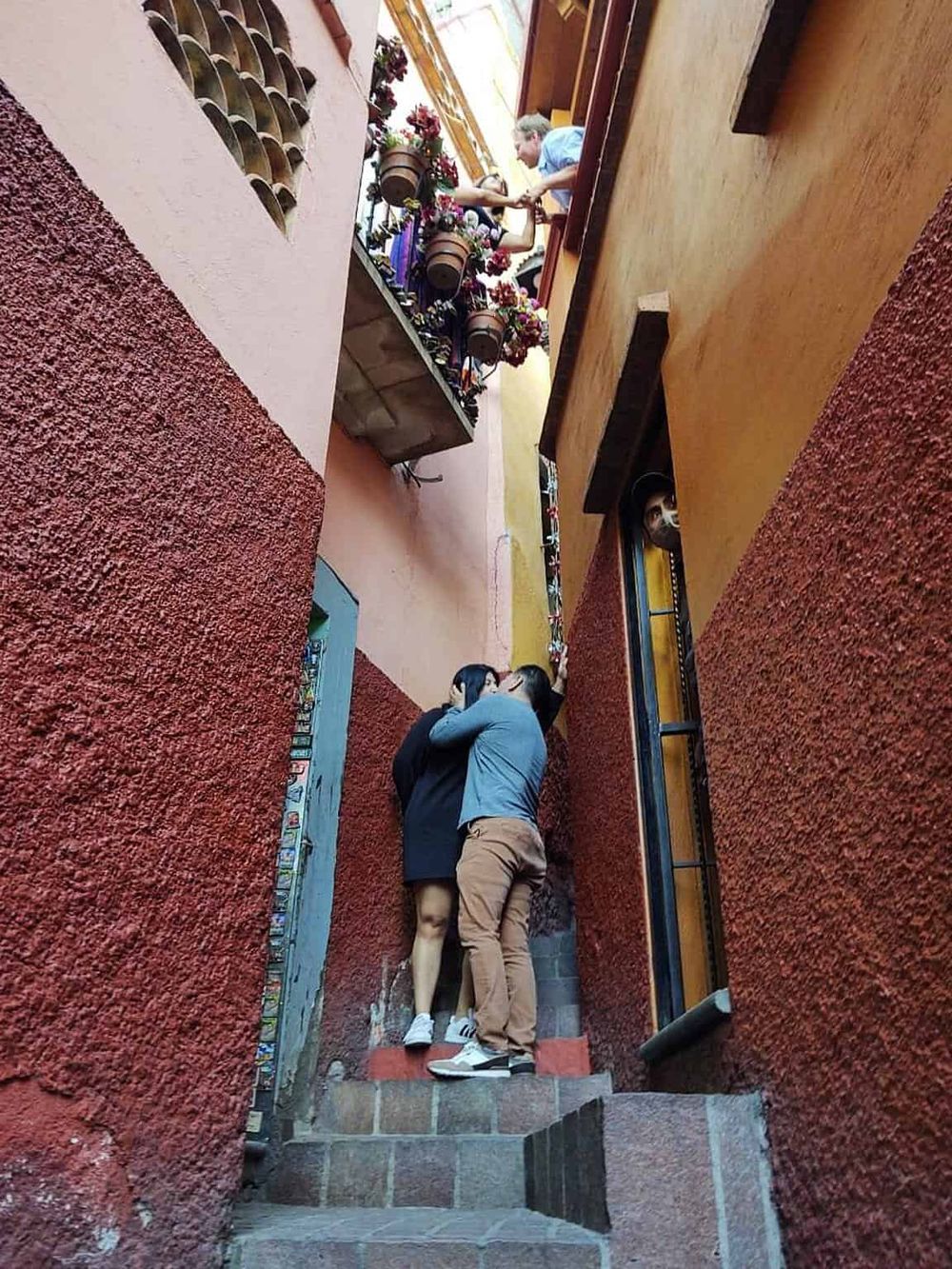 Colorful narrow alley with two people kissing and a couple on a balcony, vibrant Mexican city scene.