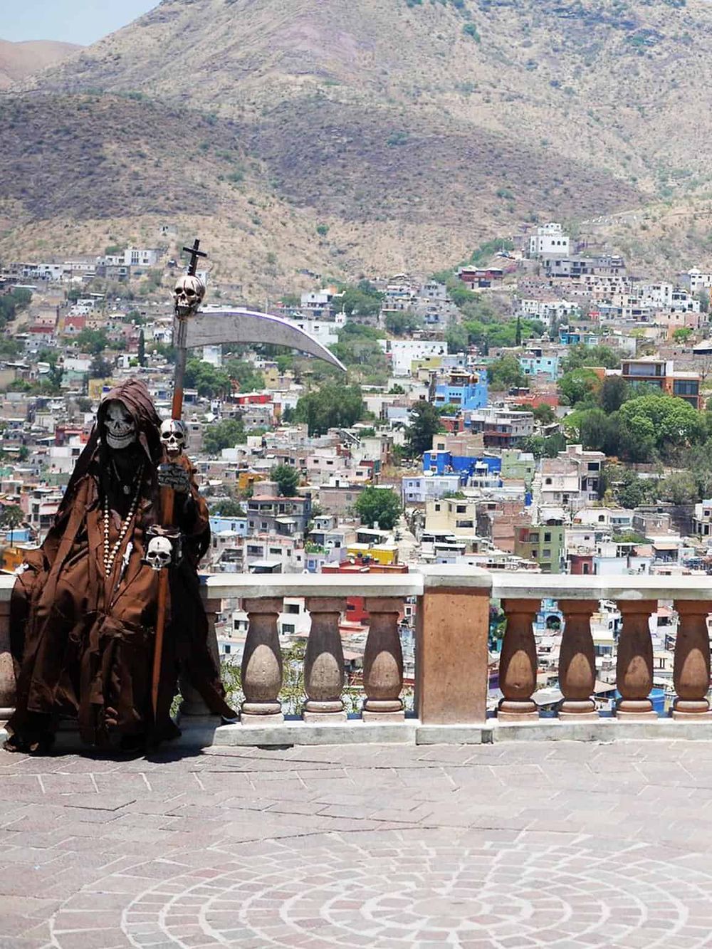 Skull-themed statue overlooking a colorful hillside cityscape in Mexico, blending culture and history.