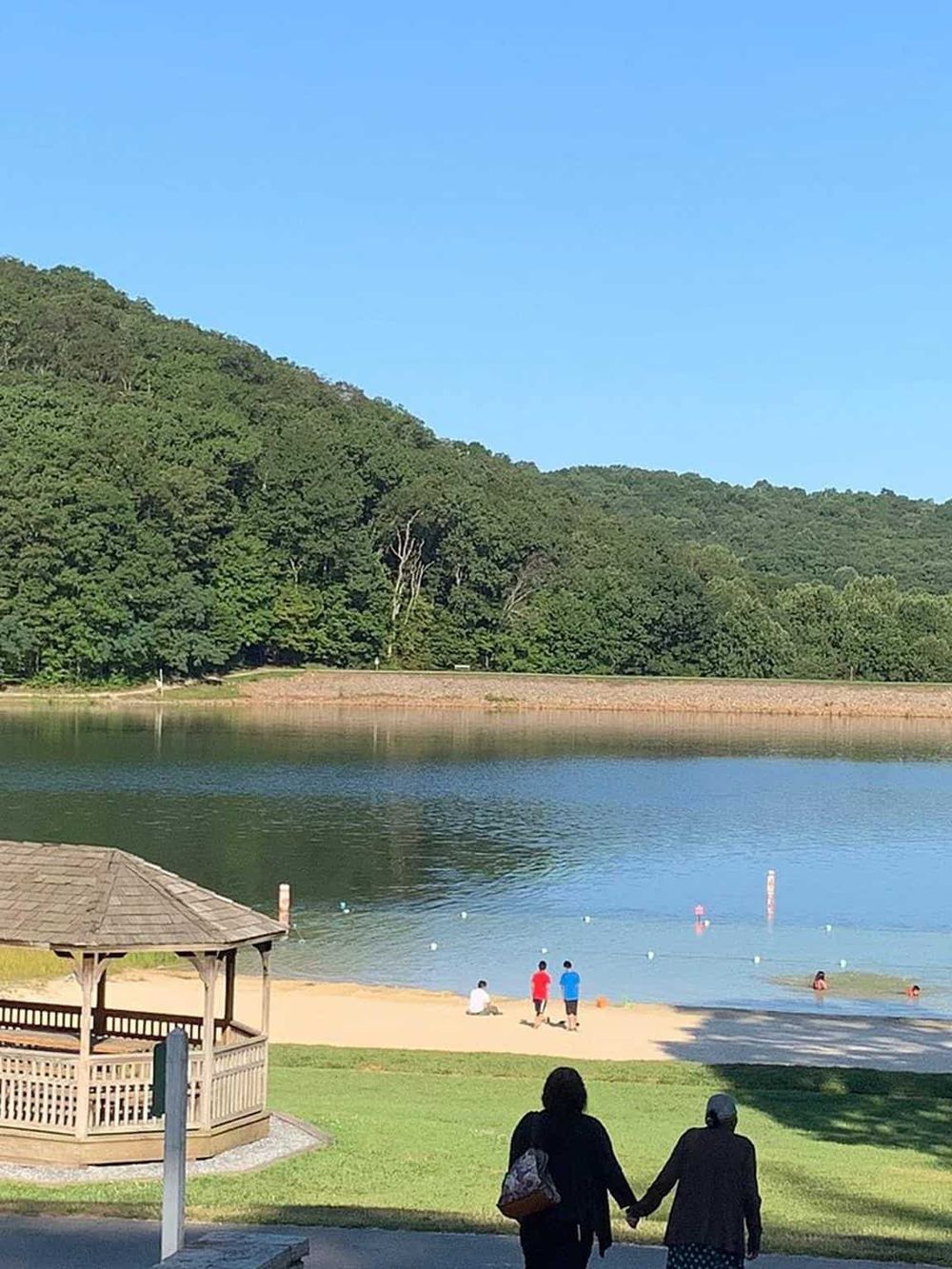 Serene lake with green hills, featuring a sandy beach, picnic shelter, and people enjoying outdoor leisure activities.