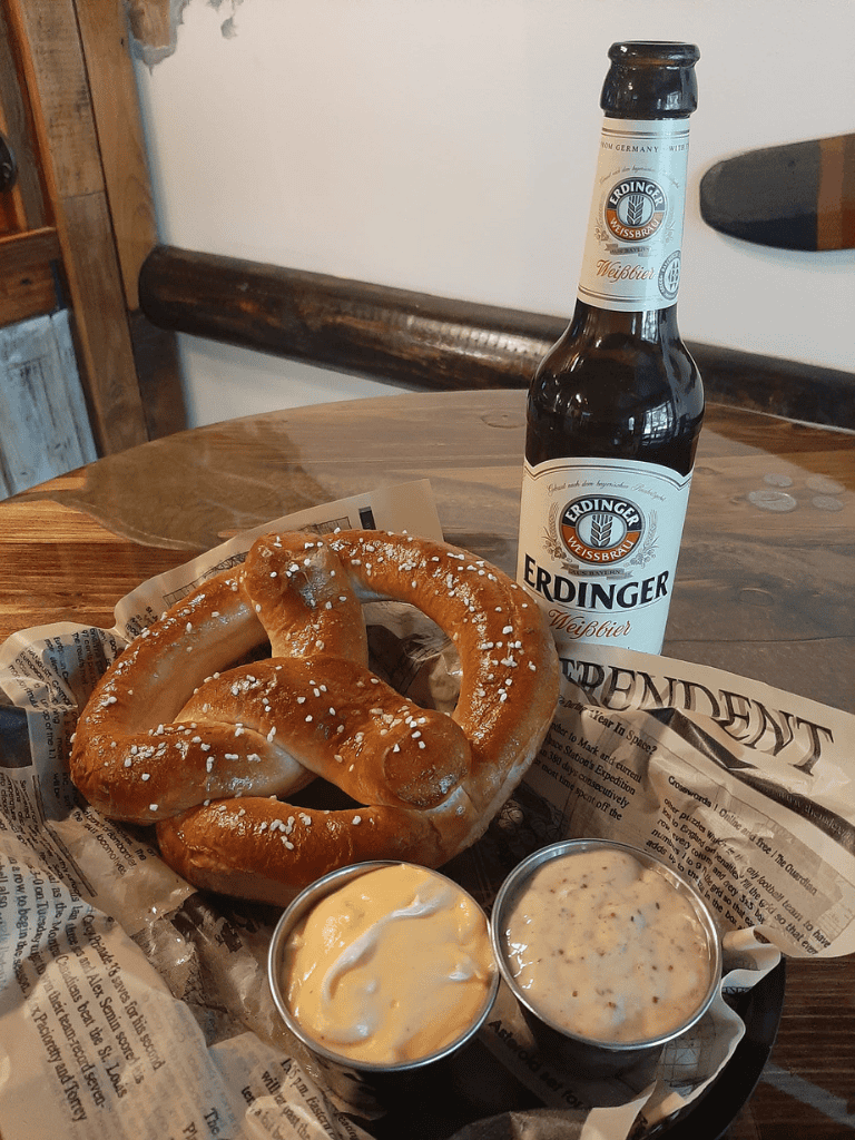 Golden pretzel with salt, Erdinger Weissbier beer, creamy cheese, and mustard on rustic wooden table.